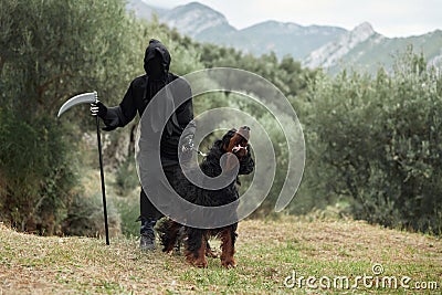 Hooded Person with Dog, Mysterious cloaked figure stands in a natural setting, holding a scythe Stock Photo