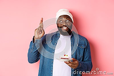 Hopeful Black guy celebrating birthday, making wish with fingers crossed, holding bday cake with candle, standing Stock Photo