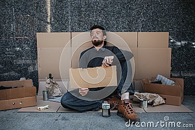 Hopeless and homeless person is sitting on cardboard on the ground and holding a piece of cardboard. He looks tired and Stock Photo