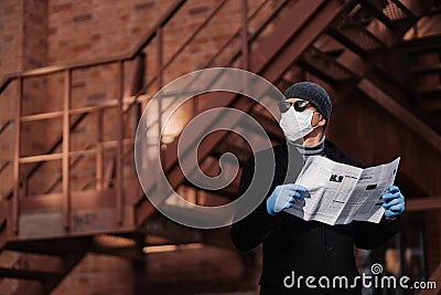 Horiontal shot of pensive man poses with newspaper, wears protective mask and rubber gloves, looks aside, thinks about self Stock Photo