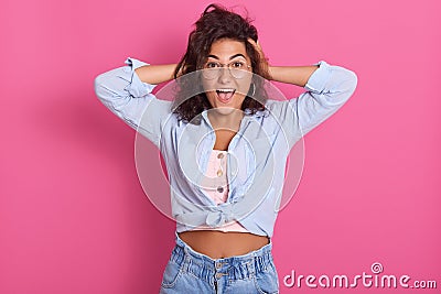 Horiontal shot of shouting, screaming young woman keeps hands on head, female dressed blue shirt and spectacles, studio portrait Stock Photo