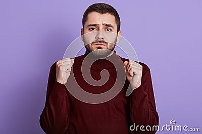 Horizontal indoor photo of hopeful energetic handsome raising hands, clenching fists, wearing dark red sweatshirt, looking Stock Photo