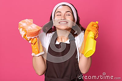 Horizontal indoor picture of delighted cheerful young female closing her eyes, raising arms, holding detergent and washcloth, Stock Photo