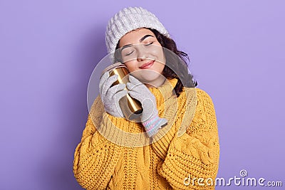 Horizontal indoor shot of tender peaceful young brunette closing eyes, smiling, holding thermomug, putting it close to face, Stock Photo