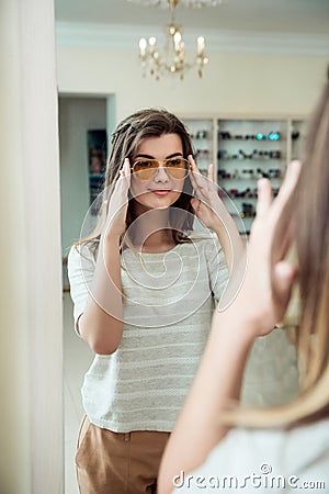 Horizontal indoor shot of trendy modern woman in casual outfit standing near mirror in optician store, trying on stylish Stock Photo