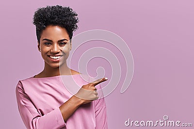 Horizontal portrait of happy dark skinned mixed race female model indicates with fore finger at blank copy space, shows Stock Photo