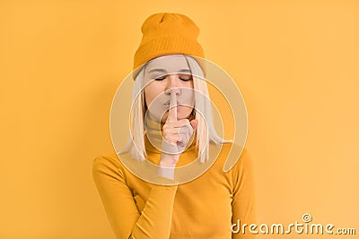 Horizontal portrait of pretty woman pressing finger against her lips, saying Shh, hush, asks to be quiet, wears in yellow clothes Stock Photo