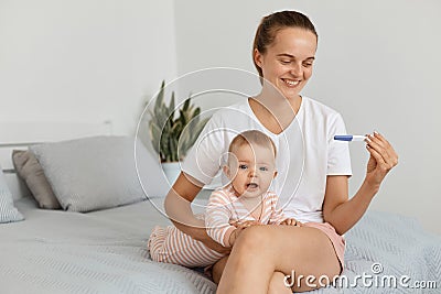 Horizontal shot of attractive female with pigtail holding pregnancy test in hands, sitting on bed in bedroom with her infant Stock Photo