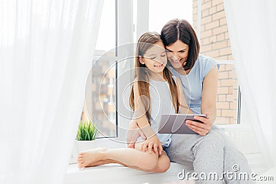 Horizontal shot of beautiful brunette mother and daughter spend free time together, sit on window sill, watch interesting movie vi Stock Photo