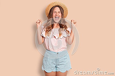 Horizontal shot of cheerful young European female clenches fists, exclaims with happiness, closes eyes, wears summer hat, blouse a Stock Photo