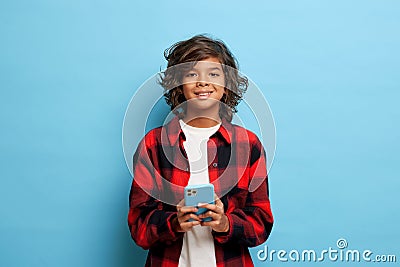 Horizontal shot of cute teenager with curly hair, holds mobile phone, waits for message, smiles gently, dressed in red Stock Photo