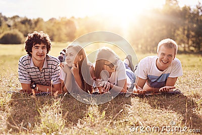 Horizontal shot of four happy friends lie on ground, have picnic during sunny summer day, have positive expressions, smile pleasan Stock Photo