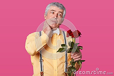 Horizontal shot of grey haired elderly man in formal fashionable clothes, keeps hand on neck, has pain, looks aside, holds red Stock Photo