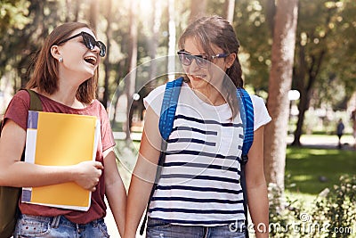 Horizontal shot of happy friends hold hand and have joyful expressions, dressed in casual clothes, hold rucksacks and books, tell Stock Photo