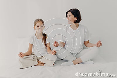 Horizontal shot of happy relaxed brunette mother and daughter pose in lotus pose, try to relax, keep legs crossed, dressed in Stock Photo