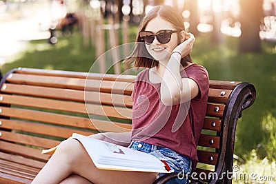 Horizontal shot of happy young female student wear shades and t shirt, reads maagzine on bench in park, has positive smile, poses Stock Photo