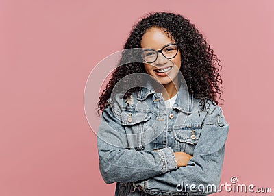 Horizontal shot of mirthful pleasant looking female model wears optical glasses and jean jacket, keeps arms folded over chest, Stock Photo