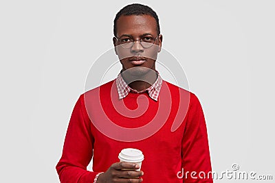 Horizontal shot of serious dark skinned man holds disposable cup of coffee, dressed in red jumper, looks confidently at Stock Photo