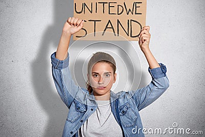 Horizontal shot of serious young woman shows fist, dressed in denim jacket, holds plate above head, being self confident in womens Stock Photo