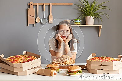Horizontal shot of upset bored Caucasian woman wearing white casual T-shirt sitting at table in kitchen, keeps hand under chin, Stock Photo