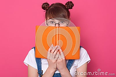 Horizontal shot of winsome young student with funny hair buns, girl hiding behind orange book, diary or planner, female wearing Stock Photo