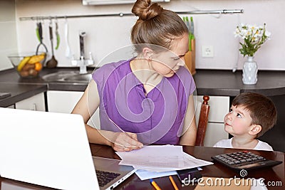 Horizontal shot of working mum being busy with documentation, makes financial report or calculates family budget, surrounded with Stock Photo