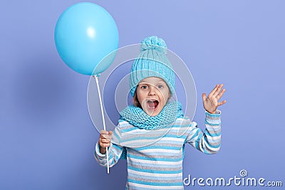 Horizontal studio picture of joyful sweet emotional little girl opening her mouth and mouth widely, raising arms, holding blue Stock Photo