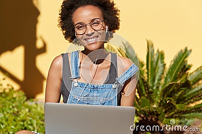 Horizontal view of black youngster with toothy smile, enjoys blogging, works on laptop computer, listens audio lesson Stock Photo