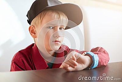 A horoizontal portrait of serious male child wearing trendy cap and red shirt having a smart watch on his wrist sitting at wooden Stock Photo