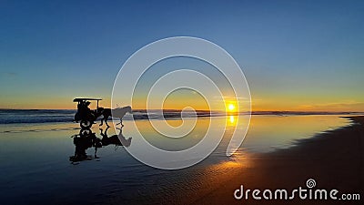 A horse-drawn carriage with a group of people driving at sunset time on the wet and reflecting sand of Parangtritis Beach in middl Stock Photo