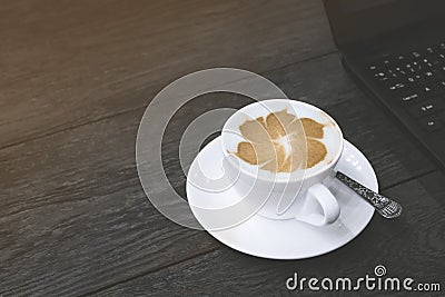 Cappuccino latte art in white ceramic cup with part of laptop computer on black wooden table Stock Photo