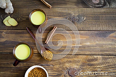 Hot Indian drink golden milk in two wooden cups on a background.Next to it are the ingredients ginger, turmeric on a wooden spoon Stock Photo