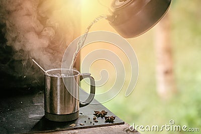 Hot tea is poured from a teapot into a metal mug with steam on a background of grass. Stock Photo