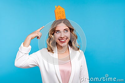 House property. Portrait of glad woman pointing at small paper house on her head. studio shot, blue background Stock Photo