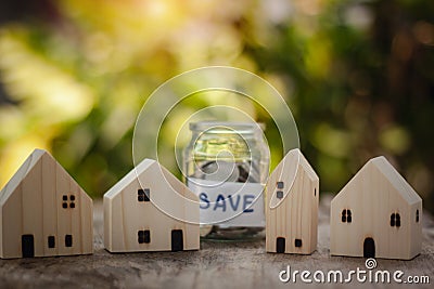 Houses model on wooden table with blurred coins in jar and sunlight bokeh background. Stock Photo