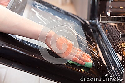 Housework and housekeeping concept. Scrubbing the stove and oven. Close up of female hand with green sponge cleaning the glass doo Stock Photo