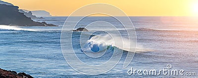 huge beautiful wave is breaking at the coastline while a breeze blows the spit water out of the sea at a wonderful sunset with Stock Photo