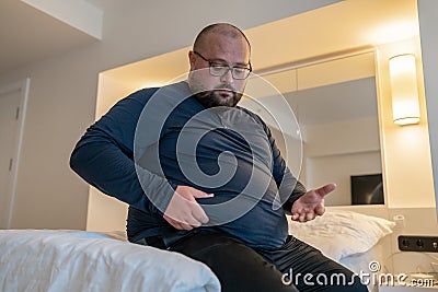 Huge man suffering from extra weight touching stomach sitting on bed in bedroom. Stock Photo