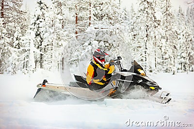 Human in the outfit of a racer in a multi-color jumpsuit and a helmet, driving a snowmobile by the deep snow surface on background Stock Photo