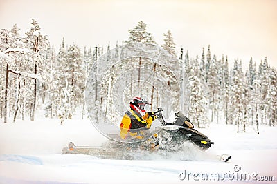 Human in the outfit of a racer in a multi-color jumpsuit and a helmet, driving a snowmobile by the deep snow surface on background Stock Photo