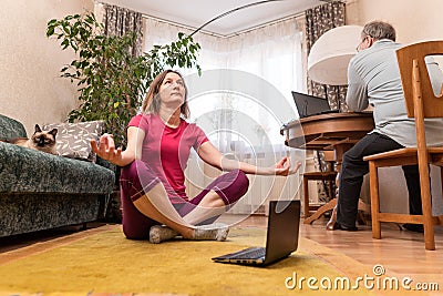 A mature woman performs yoga asana while a man works at a computer at home and cat sits on a sofa Stock Photo
