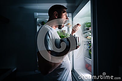 Hungry man eating food at night from open fridge. Man taking midnight snack from refrigerator Stock Photo