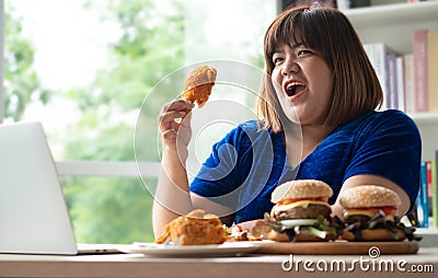 Hungry overweight woman holding Fried Chicken, hamburger on a wooden plate and Pizza on table, During work from home, gain weight Stock Photo