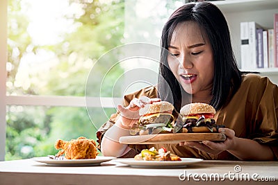 Hungry overweight woman holding hamburger on wooden plate after delivery man delivers foods at home. Concept of binge eating Stock Photo