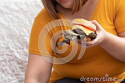 Hungry overweight woman smiling and holding hamburger and sitting in the bedroom, her very happy and enjoy to eat fast food. Conce Stock Photo