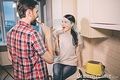 Hungry people are in kitchen. Girl sits and botes toast. Guy looks at her and does the same thing. They eat pieces of Stock Photo