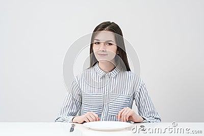 Hungry thin girl sitting at the table in front of an empty plate with a knife and fork. Stock Photo