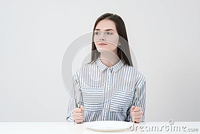 Hungry thin girl sitting at the table in front of an empty plate with a knife and fork. Stock Photo