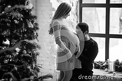 Husband kisses belly of his pregnant wife near Christmas tree. B Stock Photo