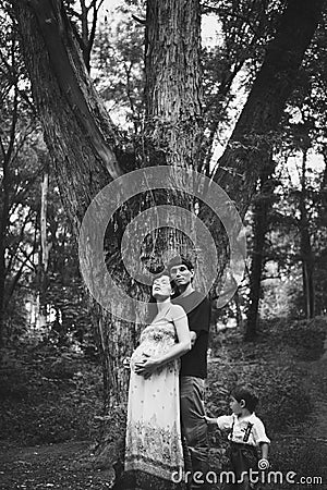 A husband, a pregnant wife and a little son are standing near a big tree, the child is holding a dad by his pants Stock Photo